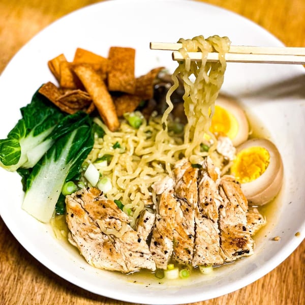 Chopsticks lifting ramen noodles from a bowl with chicken, bok choy, and soft-boiled eggs.