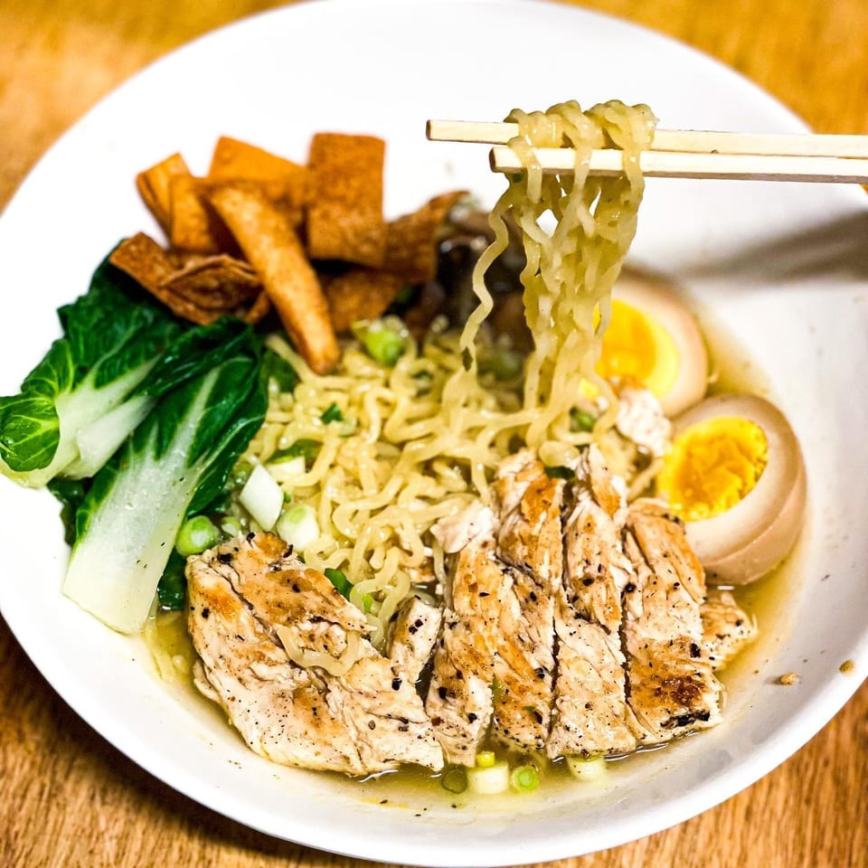 Chopsticks lifting ramen noodles from a bowl with chicken, bok choy, and soft-boiled eggs.