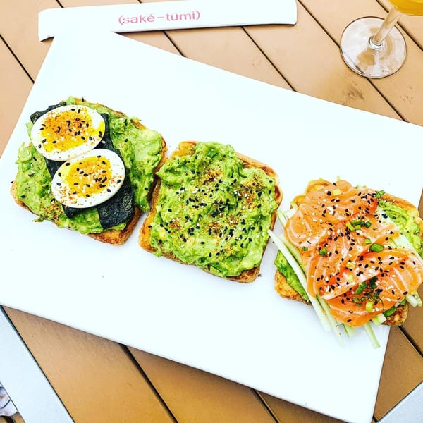 Three avocado toasts on a white plate, topped with egg, salmon, and sesame seeds.